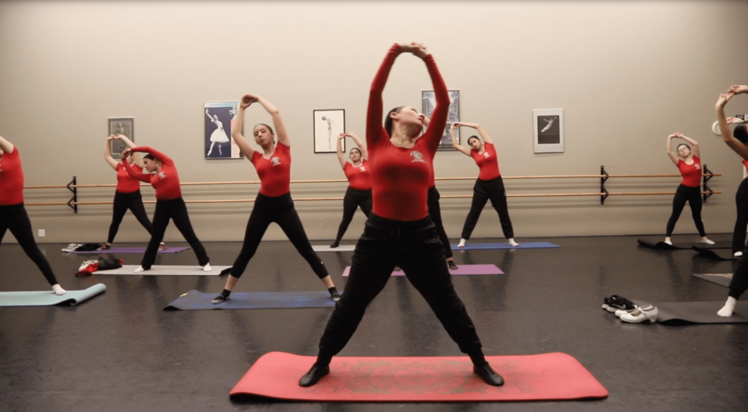 A group of dancers in red tops practice stretching exercises in a dance studio. Mirrors and ballet bars are visible in the background.