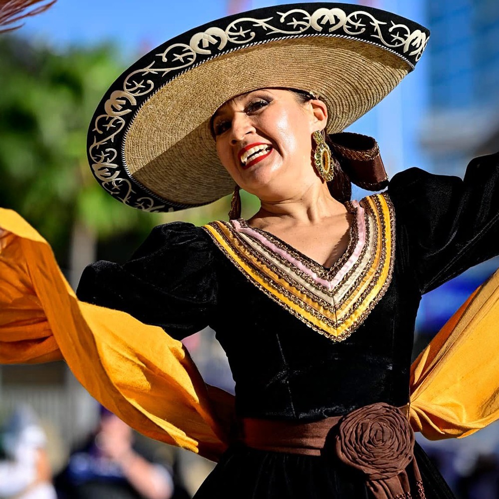 A smiling woman in traditional Mexican attire performs with vibrant yellow sleeves and a large straw hat, celebrating cultural heritage.