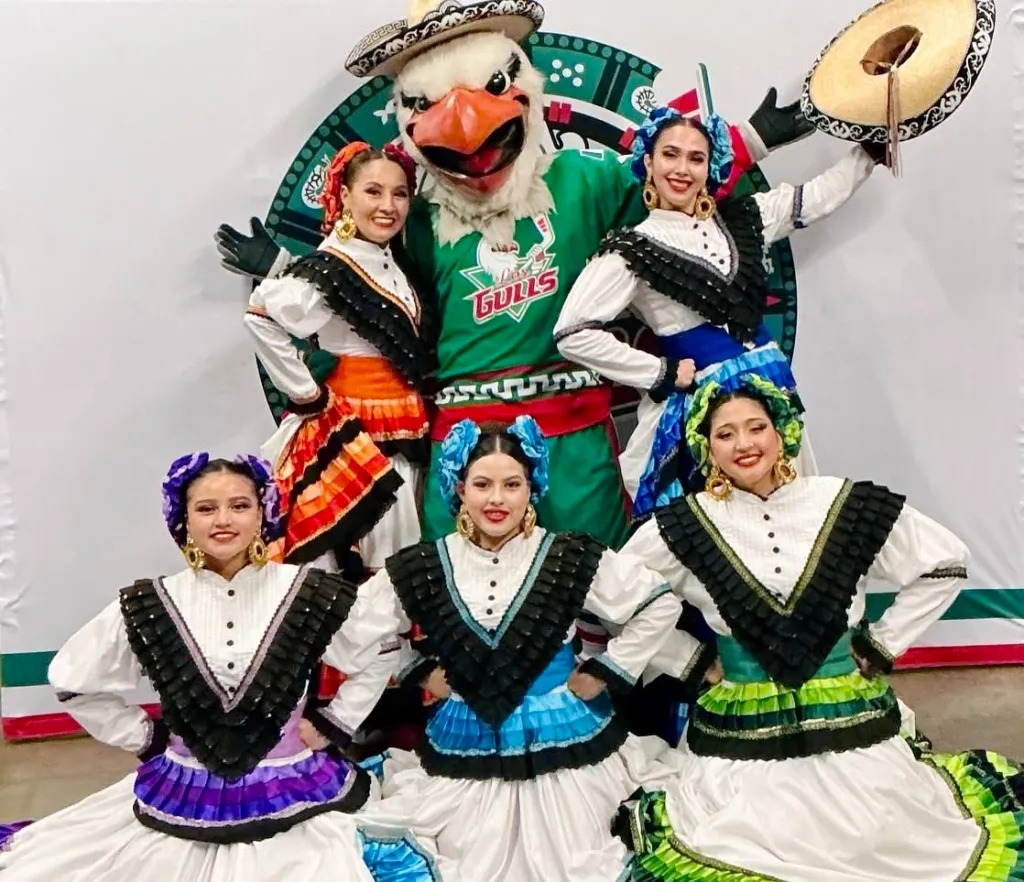 Traditional dancers in vibrant costumes pose with a team mascot at a cultural event, celebrating heritage and community spirit.