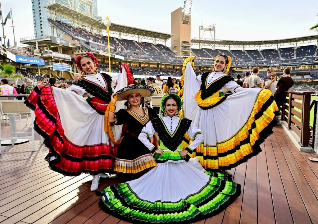 Four dancers in vibrant, traditional dresses pose on a wooden deck at a stadium, celebrating culture during an event.