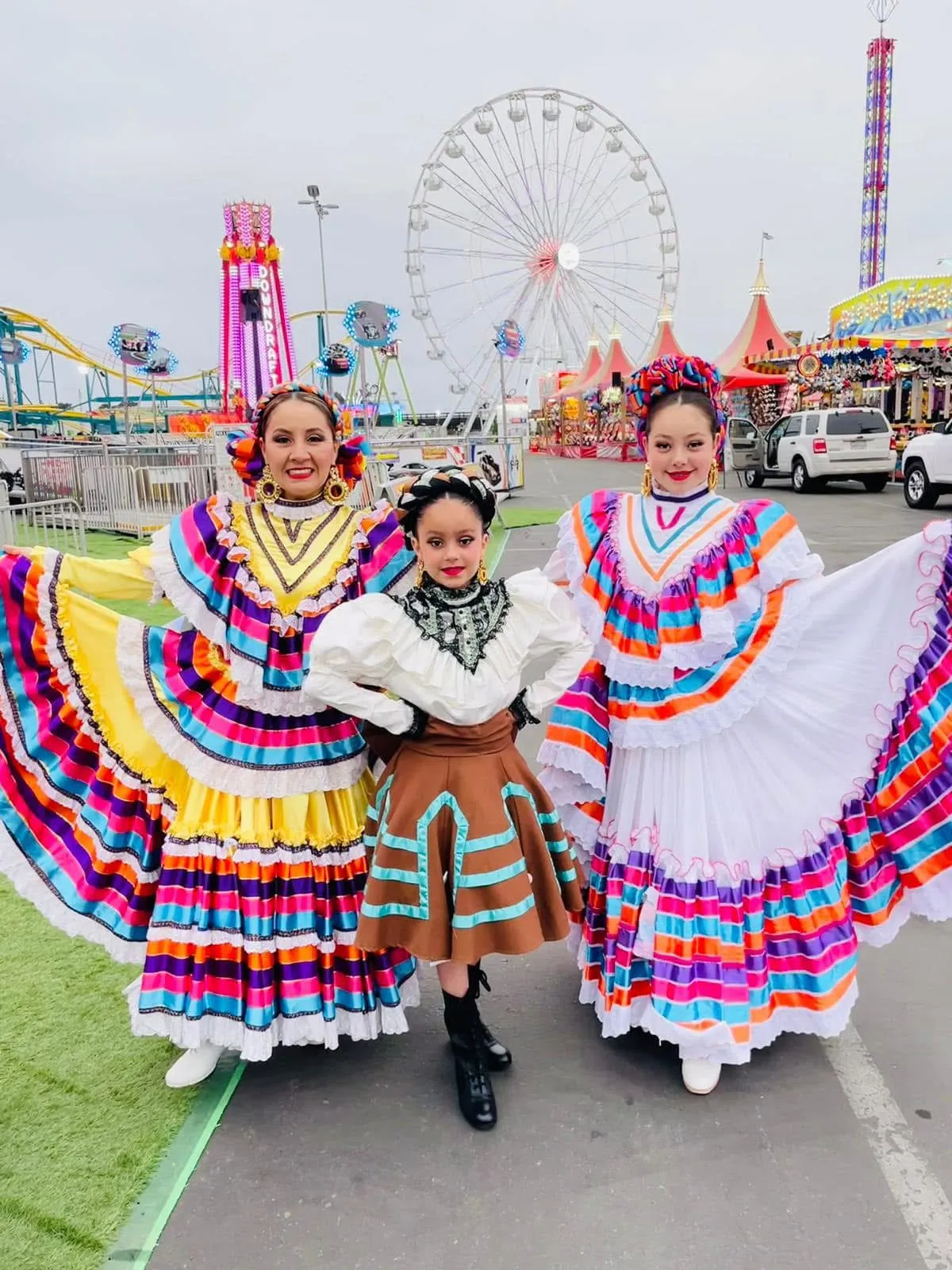 Three women in vibrant traditional dresses pose joyfully at a fairground, with a ferris wheel and colorful attractions in the background.