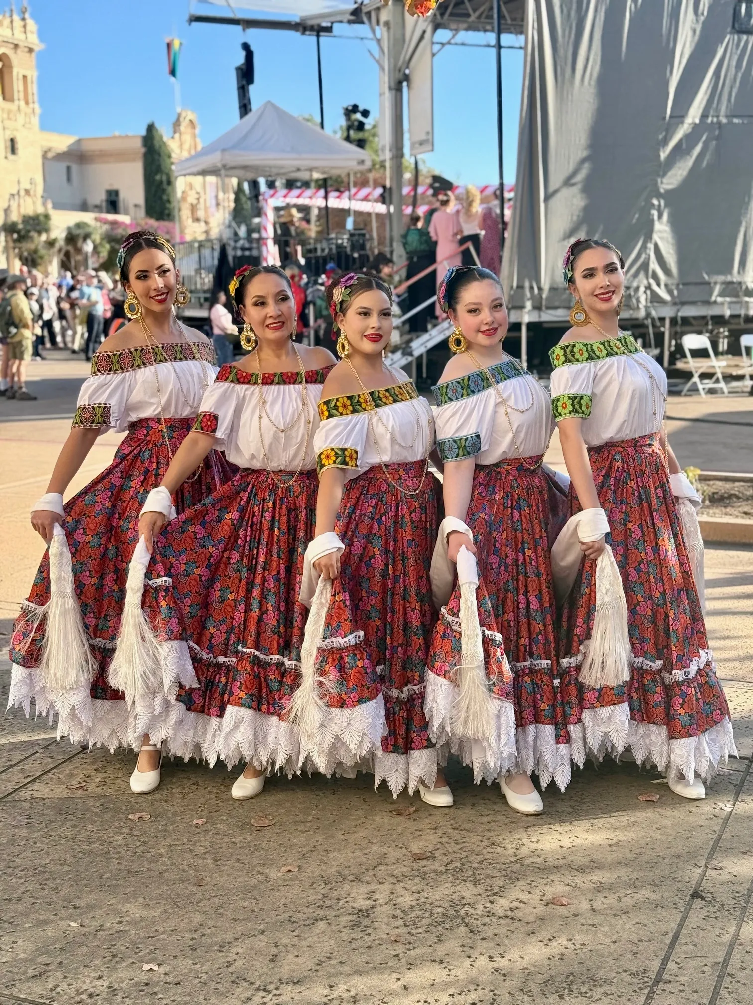 Five women in vibrant traditional dresses pose together, showcasing their colorful outfits with floral patterns, during a cultural festival.