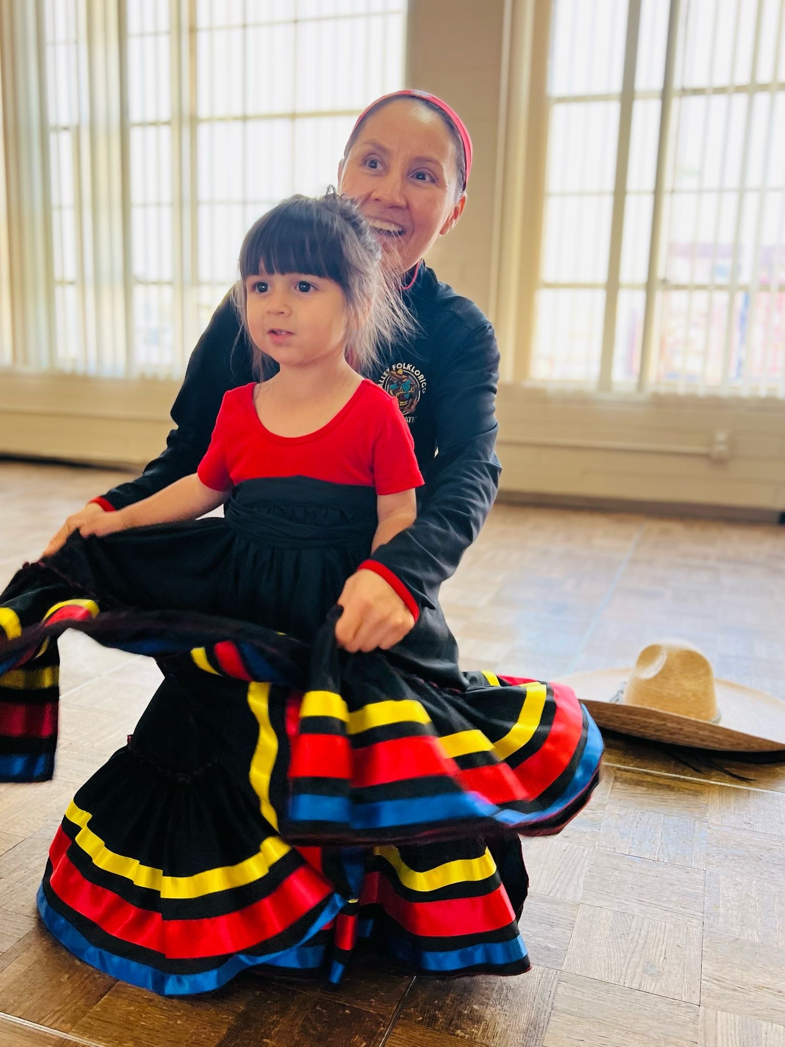 A woman joyfully dances with a young girl in traditional colorful skirts, celebrating cultural heritage in a bright, spacious room.