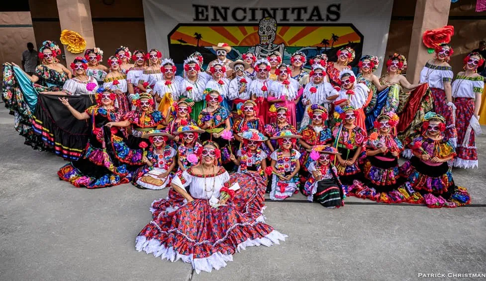 A vibrant group of dancers in colorful traditional costumes poses together, showcasing their festive makeup and expressions at a cultural event in Encinitas.