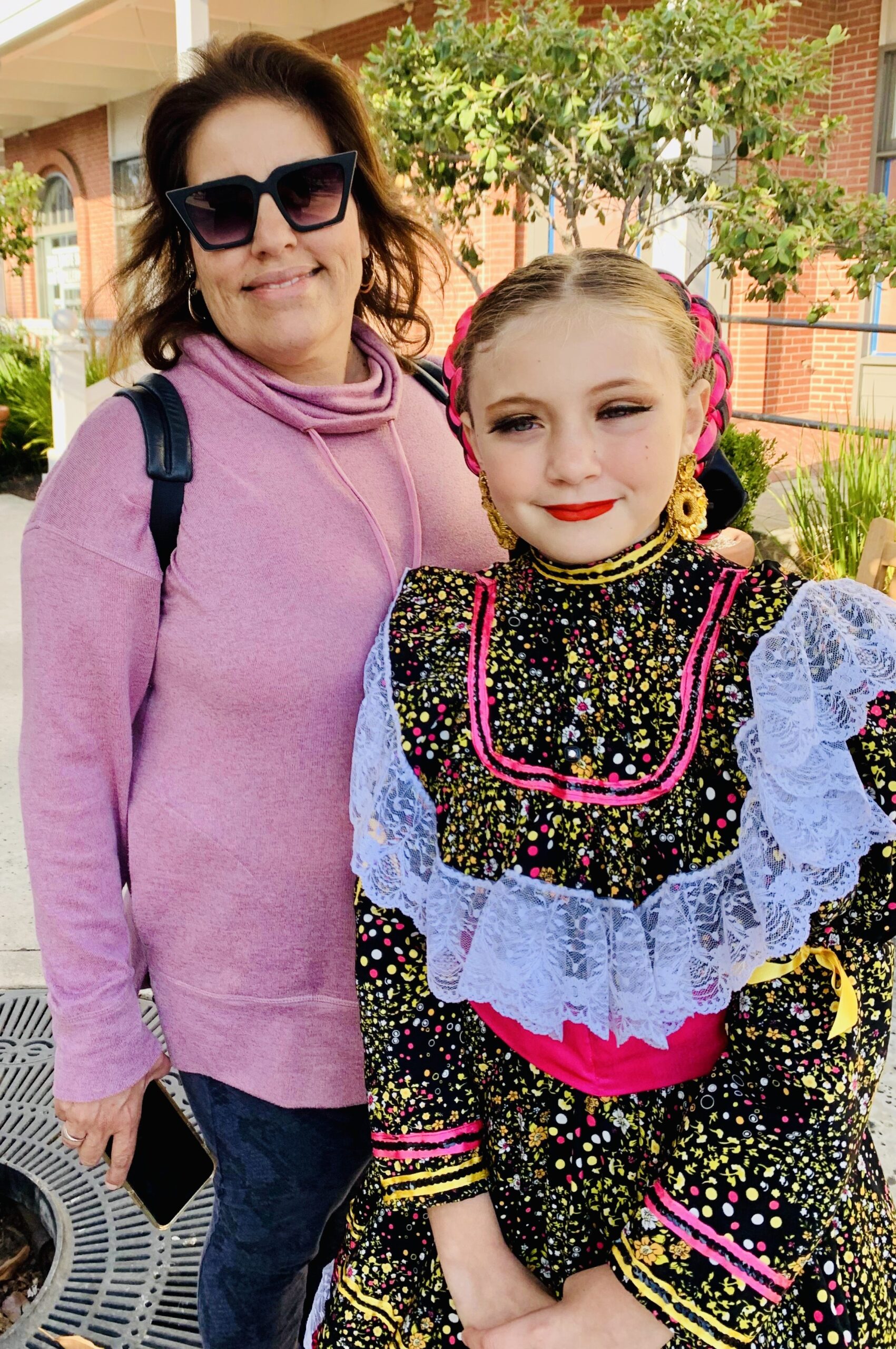 A woman in sunglasses smiles beside a girl in a colorful, traditional dress with lace details. They stand outdoors, suggesting a festive occasion.